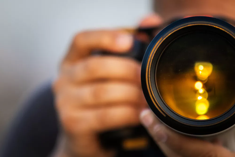 Close-up of a person's hand holding a camera, focusing on the glowing orange reflections in the lens. The blurred background adds depth and intrigue.
